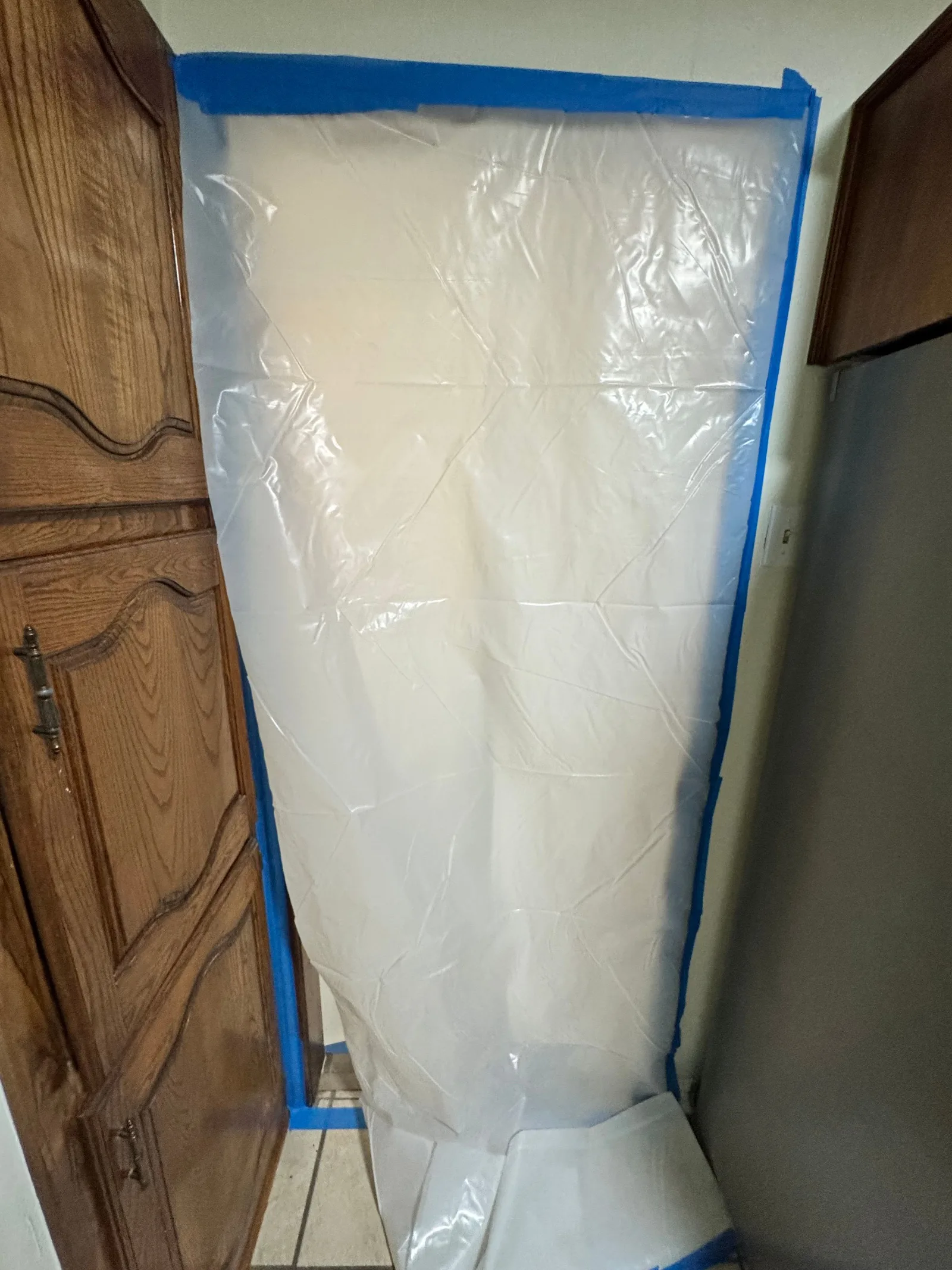 Clear plastic sheeting hangs from a room's ceiling and blue painter's tape secures it to the wall, protecting the kitchen.