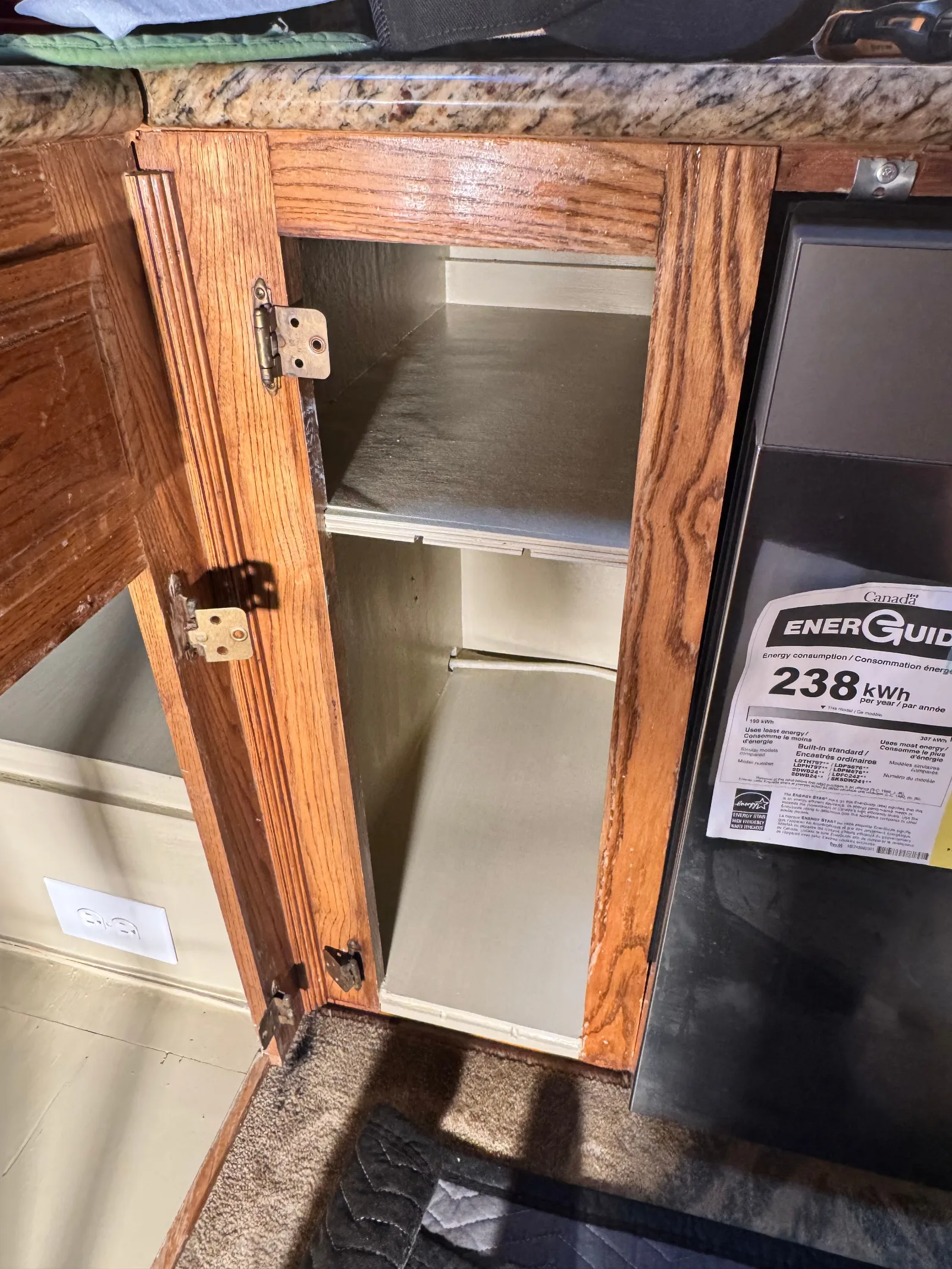 Open wooden kitchen cabinet under a granite counter next to a black dishwasher with a yellow Energy Guide label.