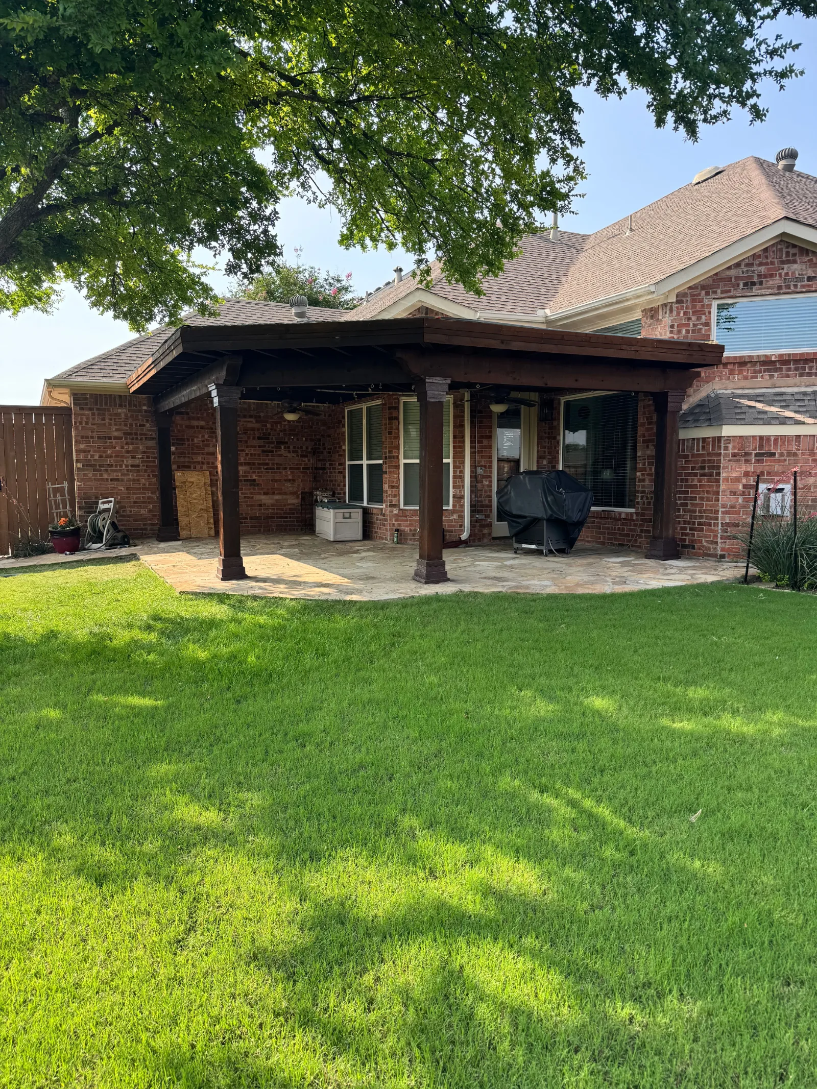 Backyard patio with a large brown wooden pergola, stone tile flooring, and a red brick house background.