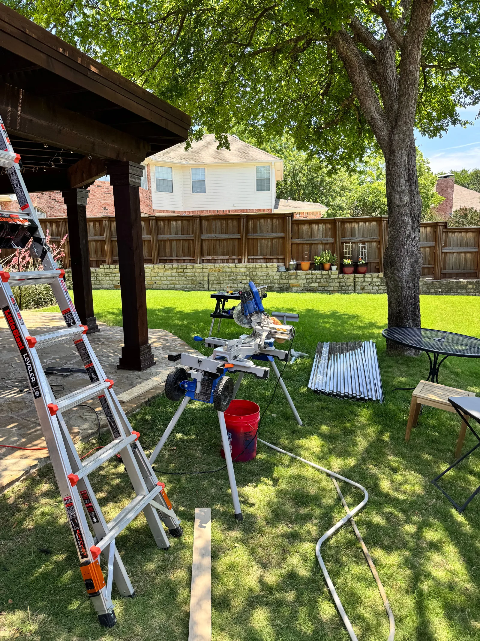 Backyard with a brown wooden pergola. A miter saw, ladder, and stack of corrugated metal panels are on green grass.