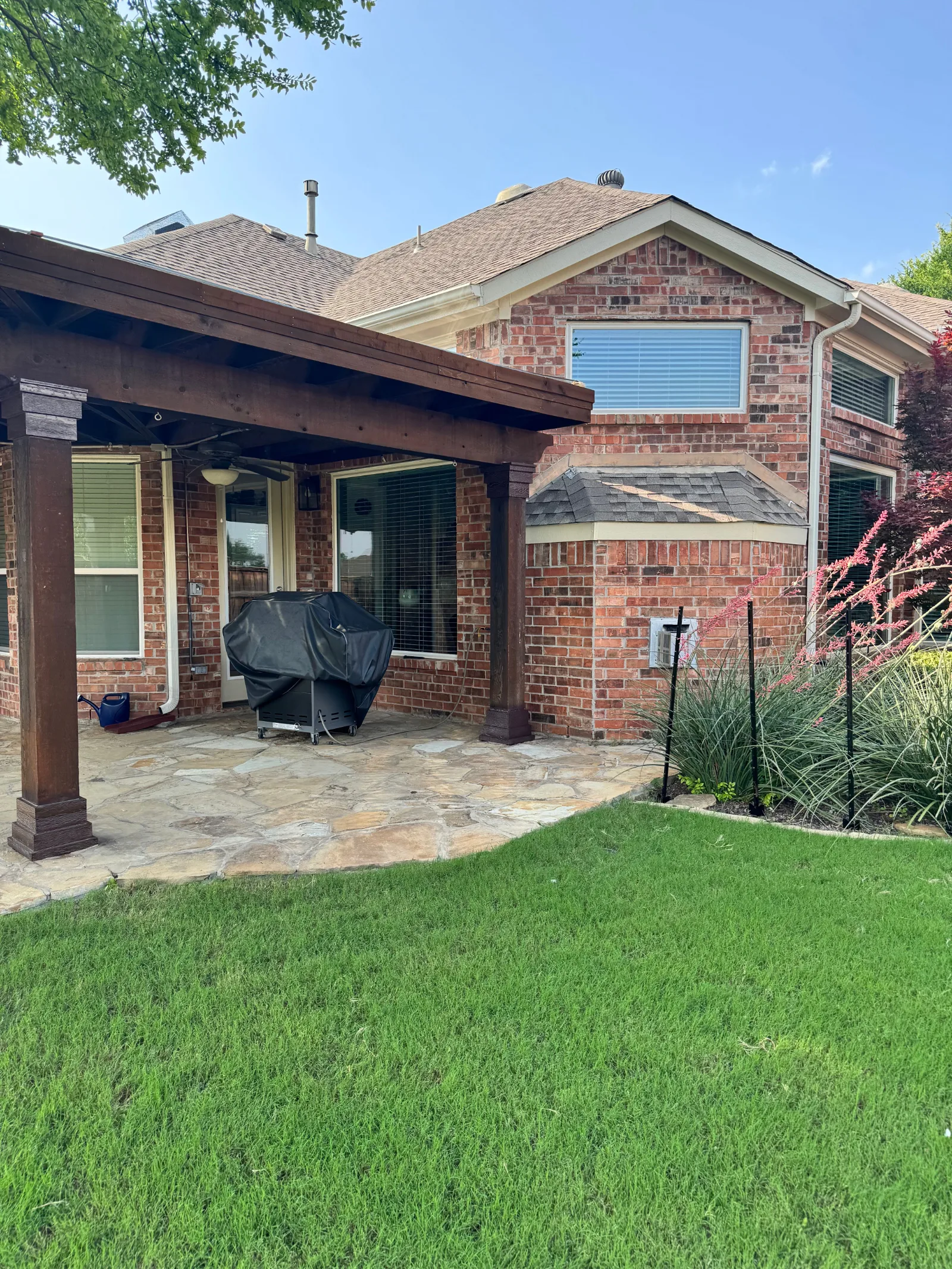 Back of a red brick house with a newly constructed dark wood pergola over a flagstone patio. Green lawn in foreground.