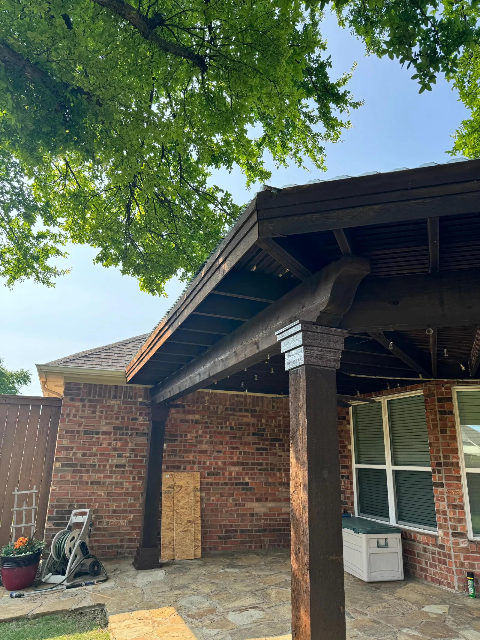 Dark wood pergola with corrugated polycarbonate roof attached to a brick home with stone patio.