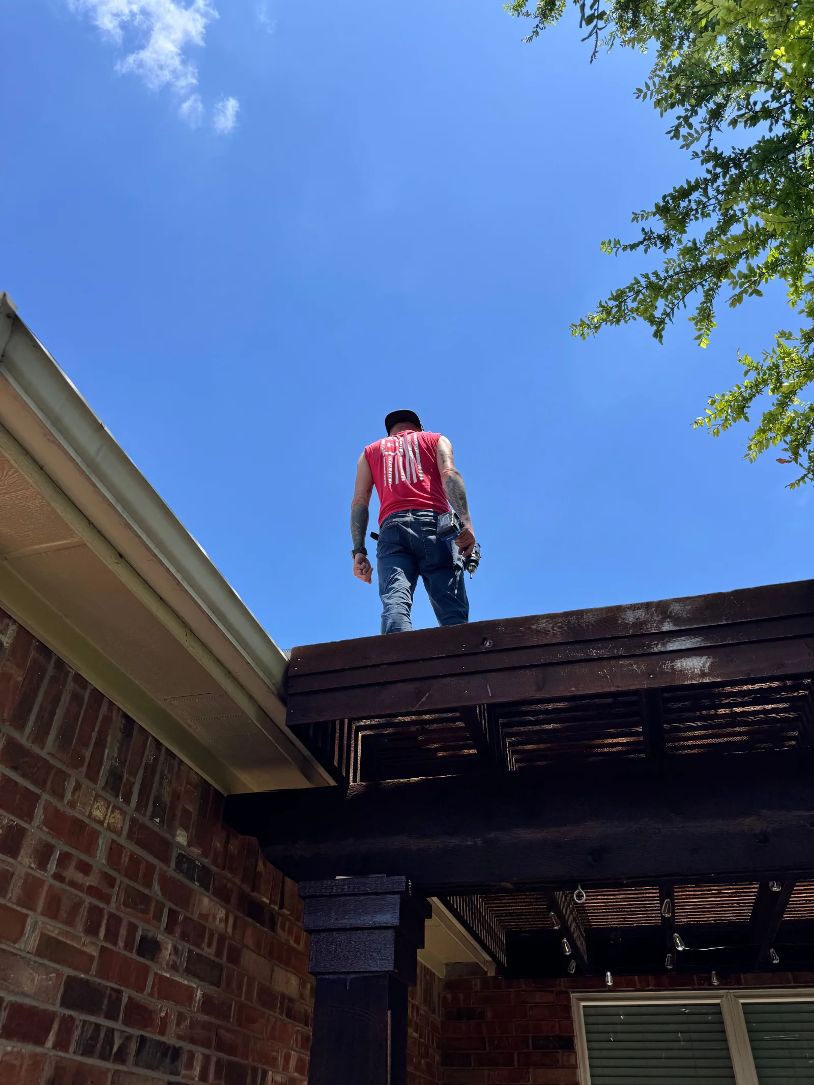 Workman in red shirt on a wooden pergola roof next to brick house under a bright blue sky.