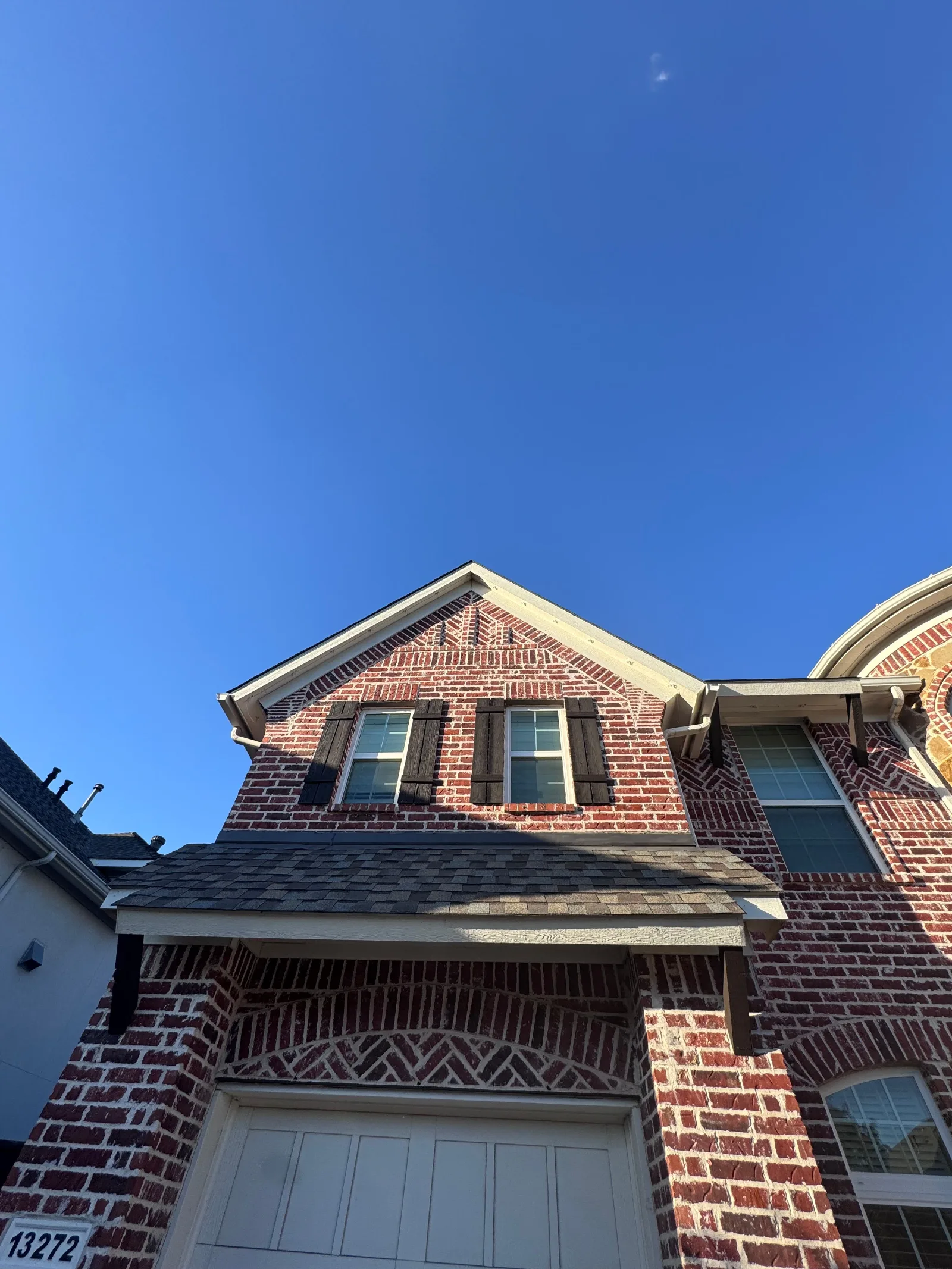 Red brick house exterior with dark shingle roof, two gabled windows with dark shutters, and a white garage door.