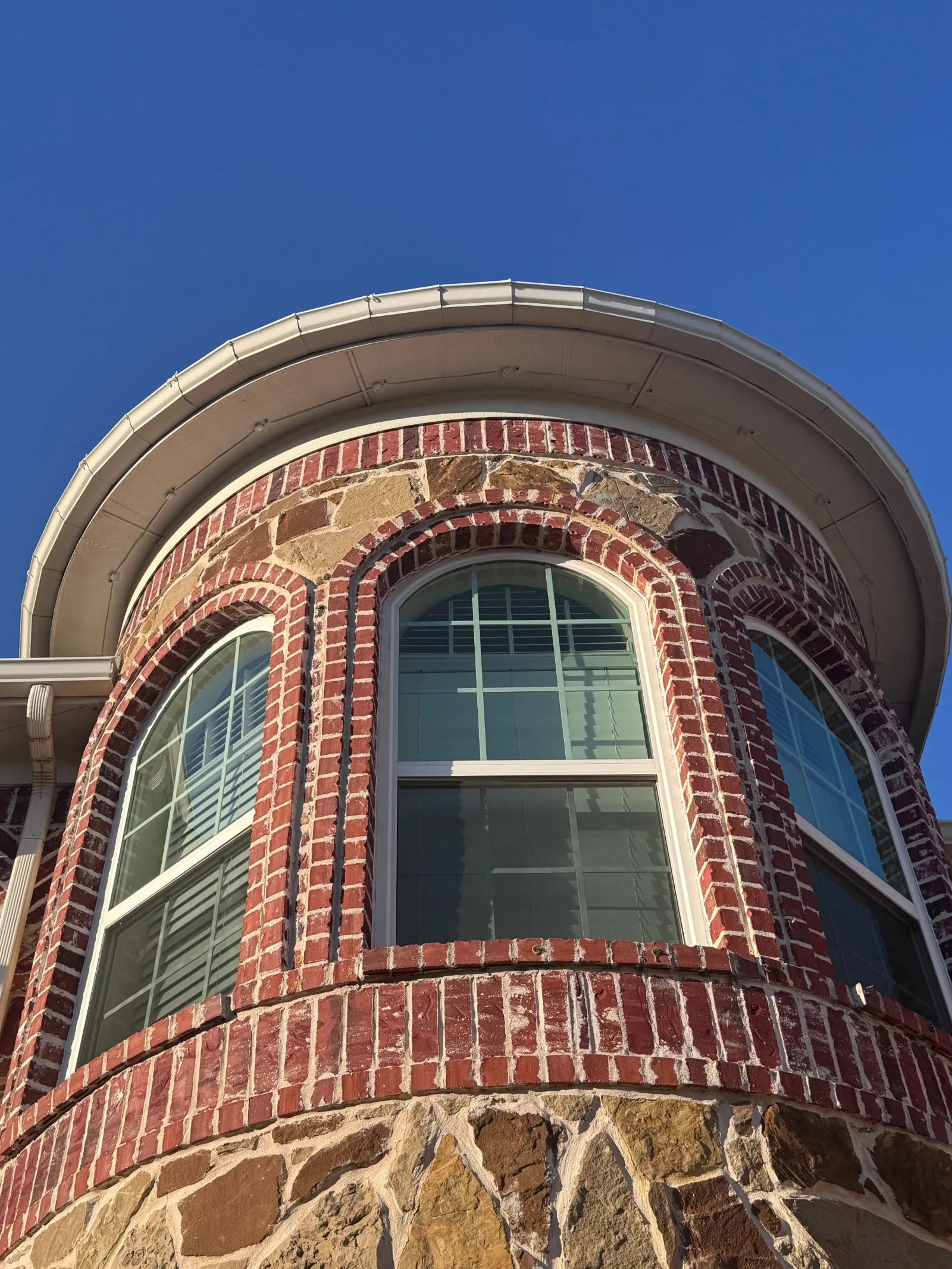 Curved red brick and stone turret of a house with arched windows and white rain gutters overhead against a blue sky.