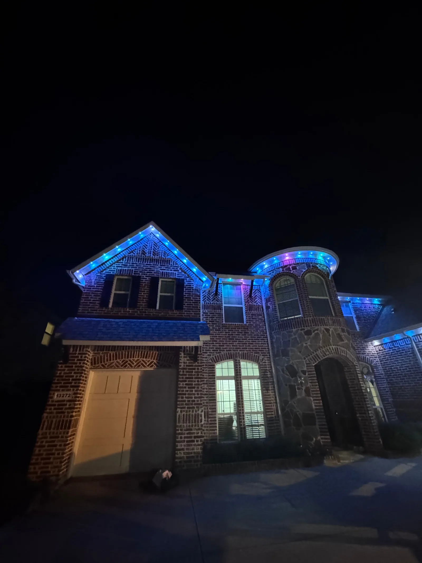 A two-story traditional brick house at night with blue and purple smart exterior lighting outlining the gables and round turr