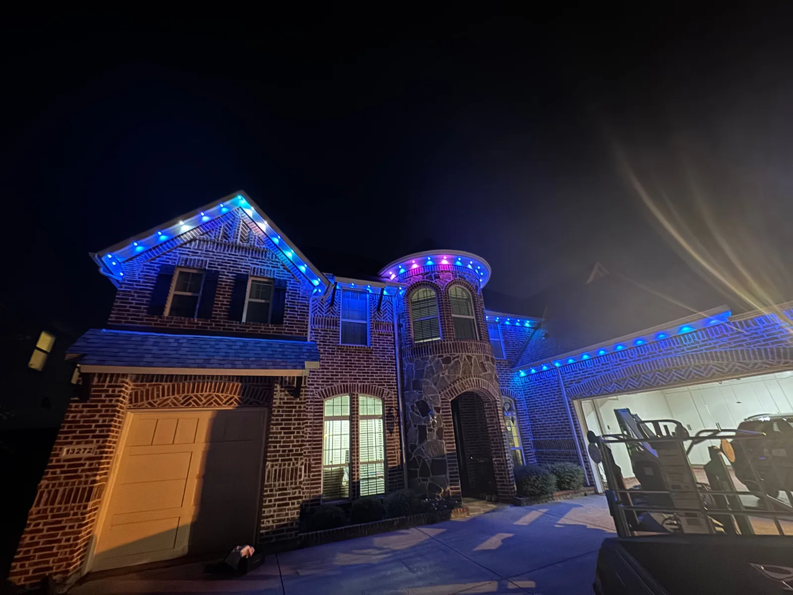 Two-story brick and stone home at night with blue and purple LED lights installed along the roofline and porch.