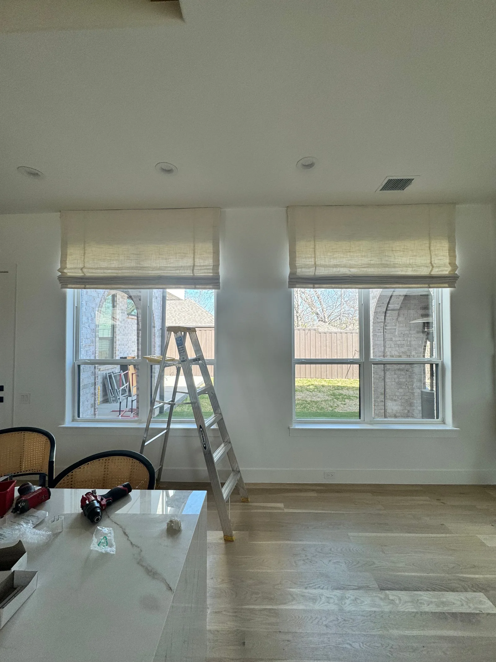 Two light-colored Roman shades installed over windows in a room with light wood floors and a kitchen island.