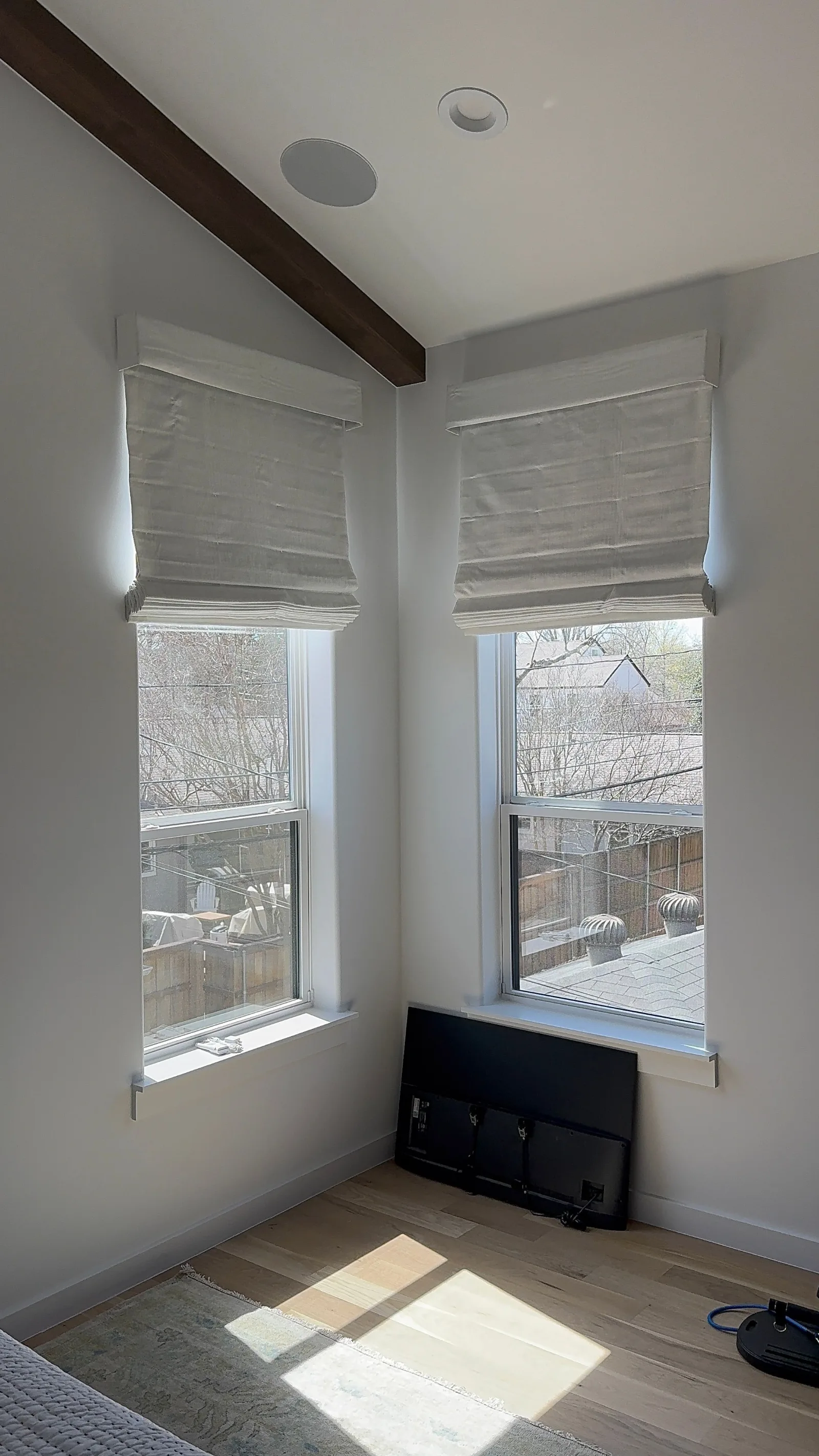 Two white Roman shades installed in a bright room with light wood floors, a decorative beam, and recessed lighting.