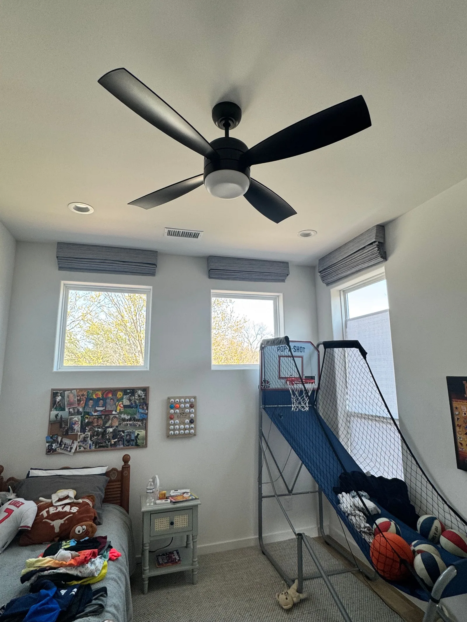 Blue-gray Roman shades in a child's bedroom with a dark wood bed frame, white walls, and a mini basketball hoop.