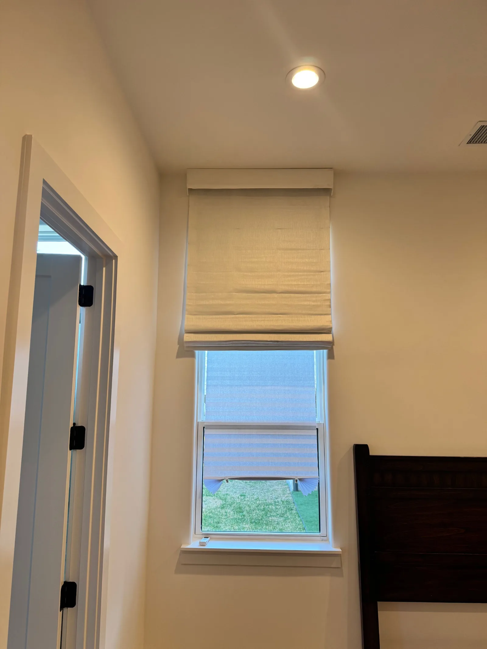 Bedroom with a tall window featuring a light-colored Roman shade over cellular shades. A dark wooden headboard is visible on