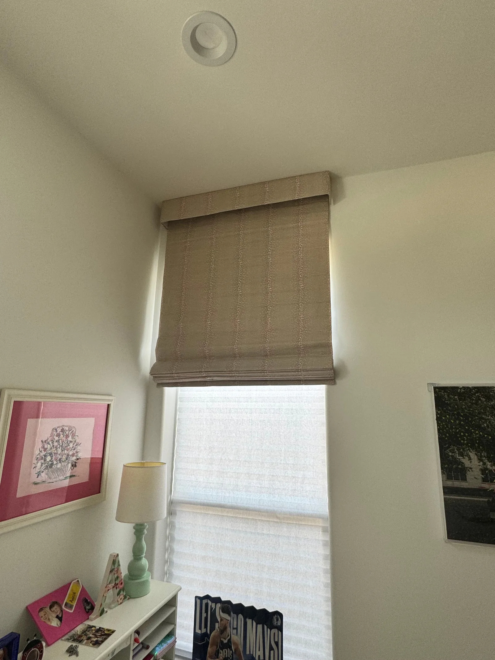 Taupe Roman shade covering a window, with a white cellular shade visible below it, in a bright room.