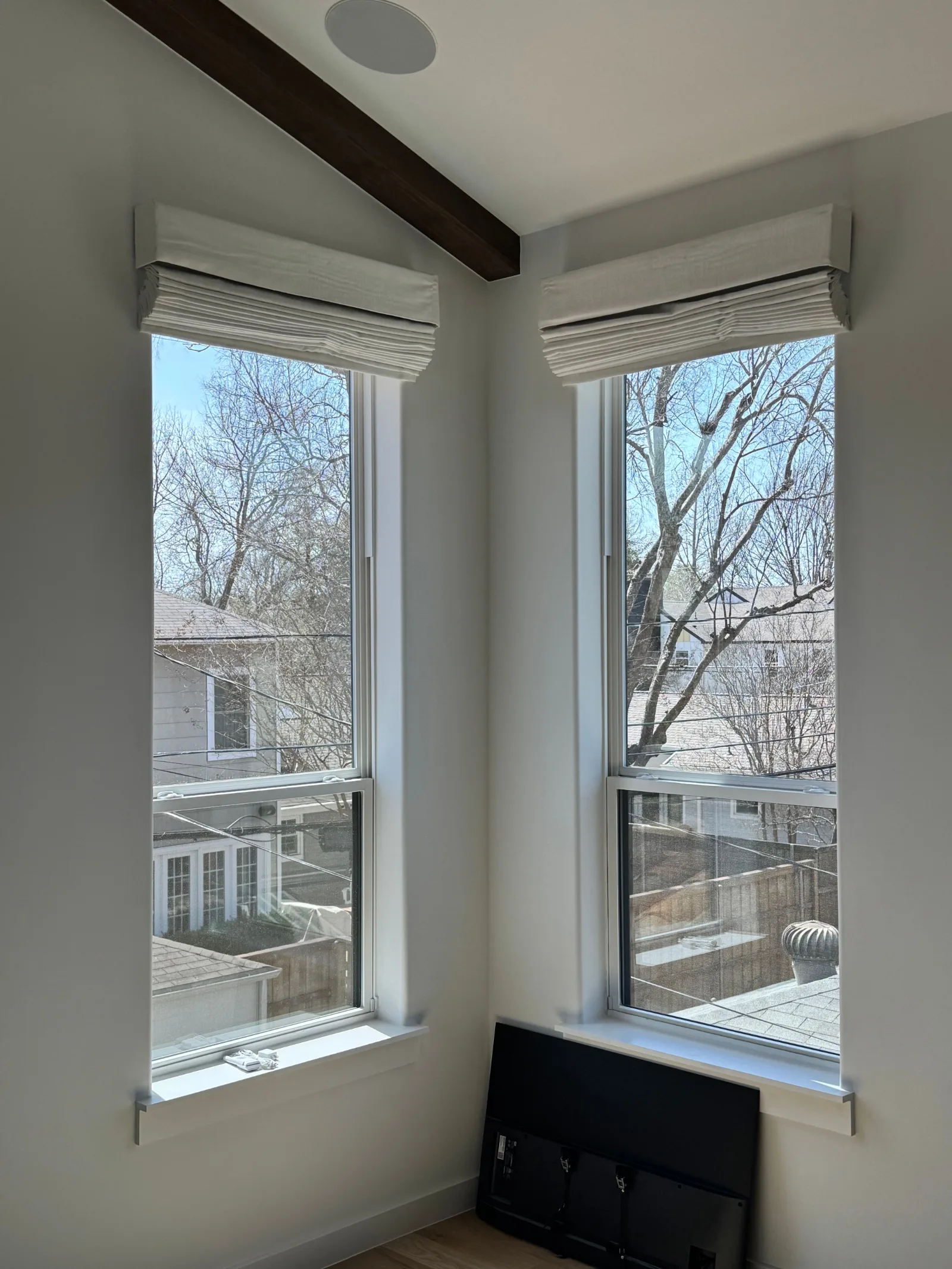 Two white Roman shades installed on adjacent windows in a room with white walls and a dark wood beam on the ceiling.