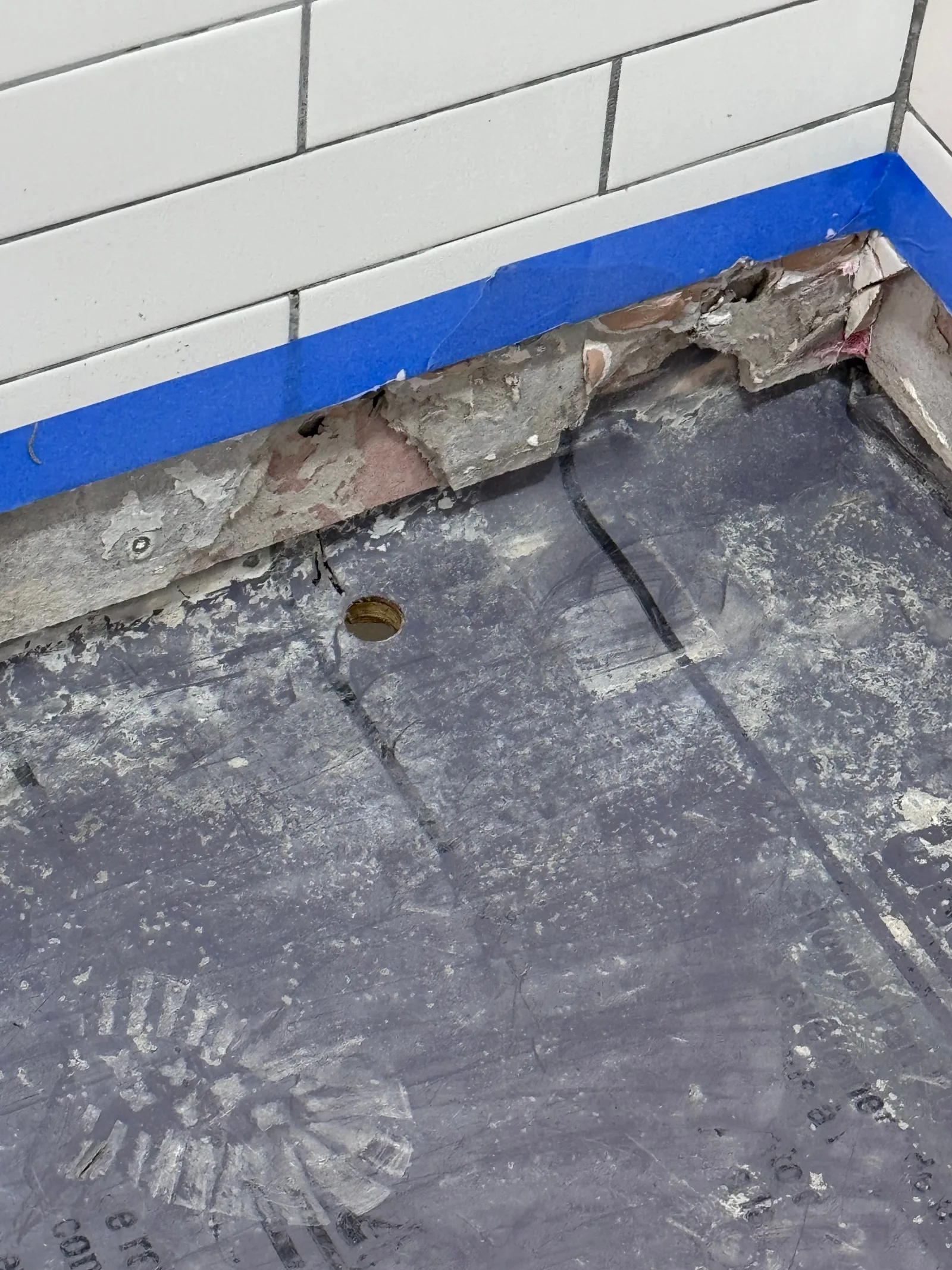 Close-up of a shower pan with a drain opening, blue painter's tape along a tiled wall, and exposed mortar.