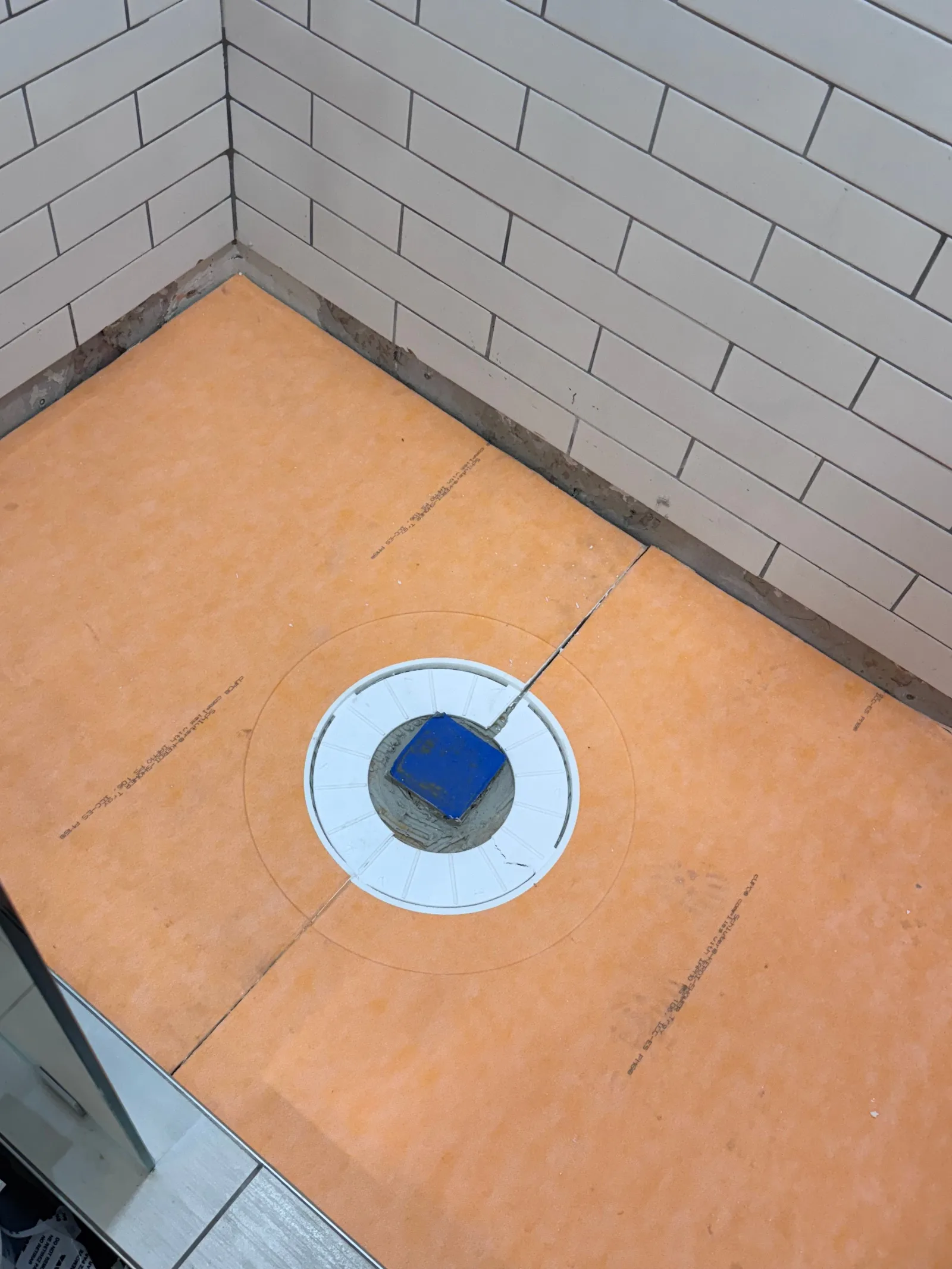Top-down view of an orange Schluter shower pan base with a white drain, adjacent to partially tiled white subway walls.