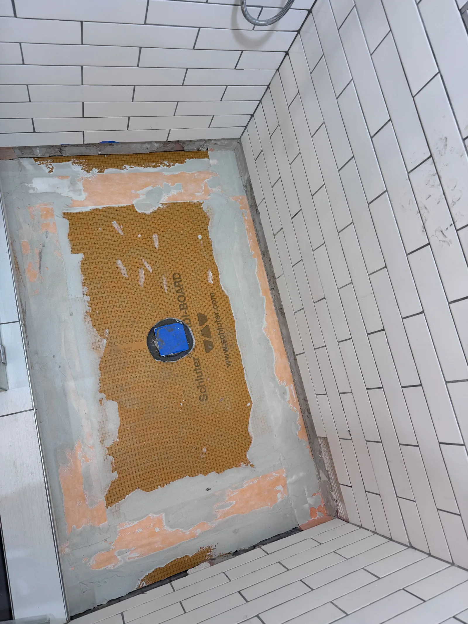 A shower pan with Schluter orange waterproofing membrane partially covered with grey mortar and white subway tile walls.