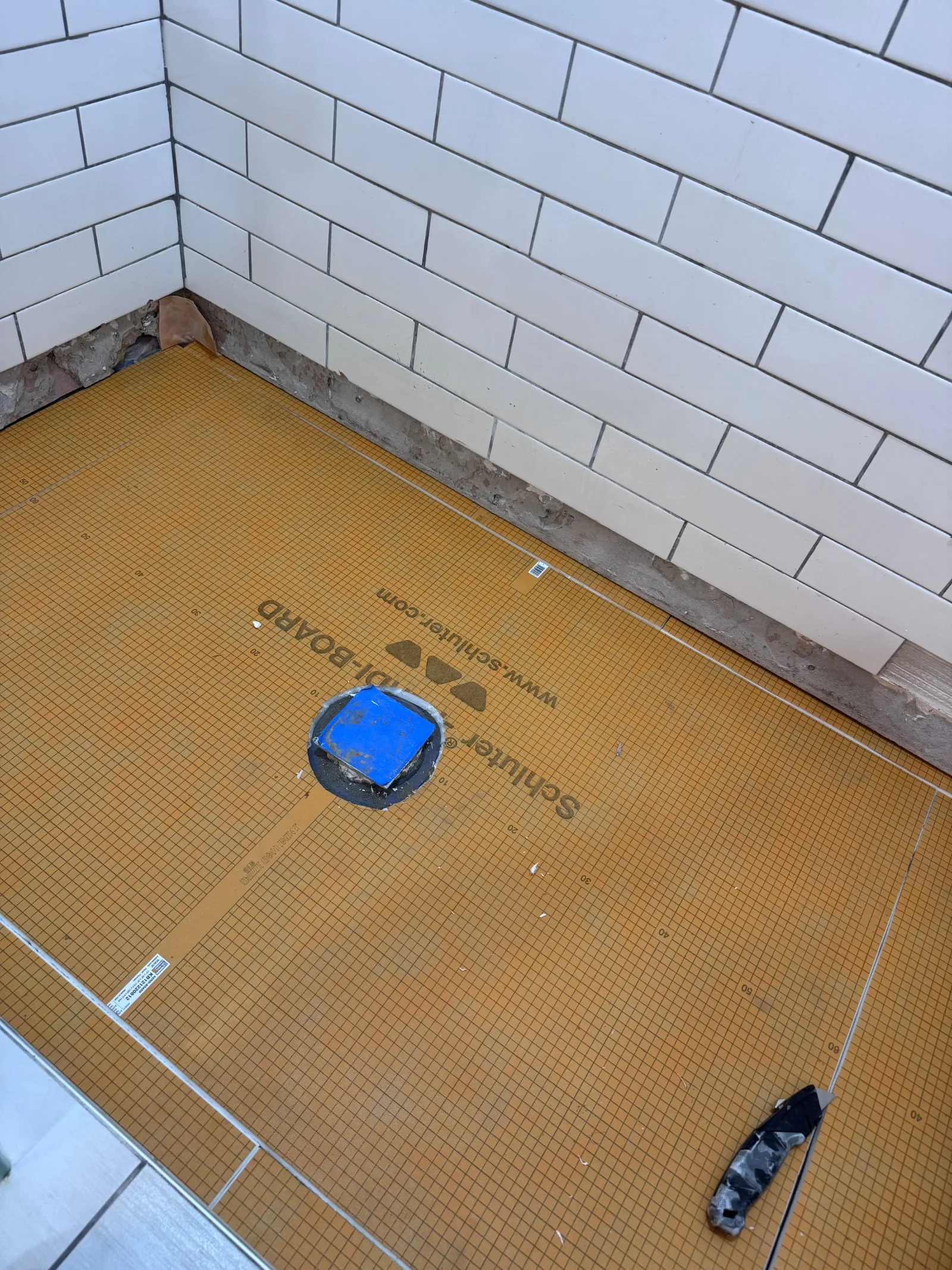 Shower floor with an orange Schluter-DITRA waterproofing membrane, partially tiled white subway walls, and a blue drain cover