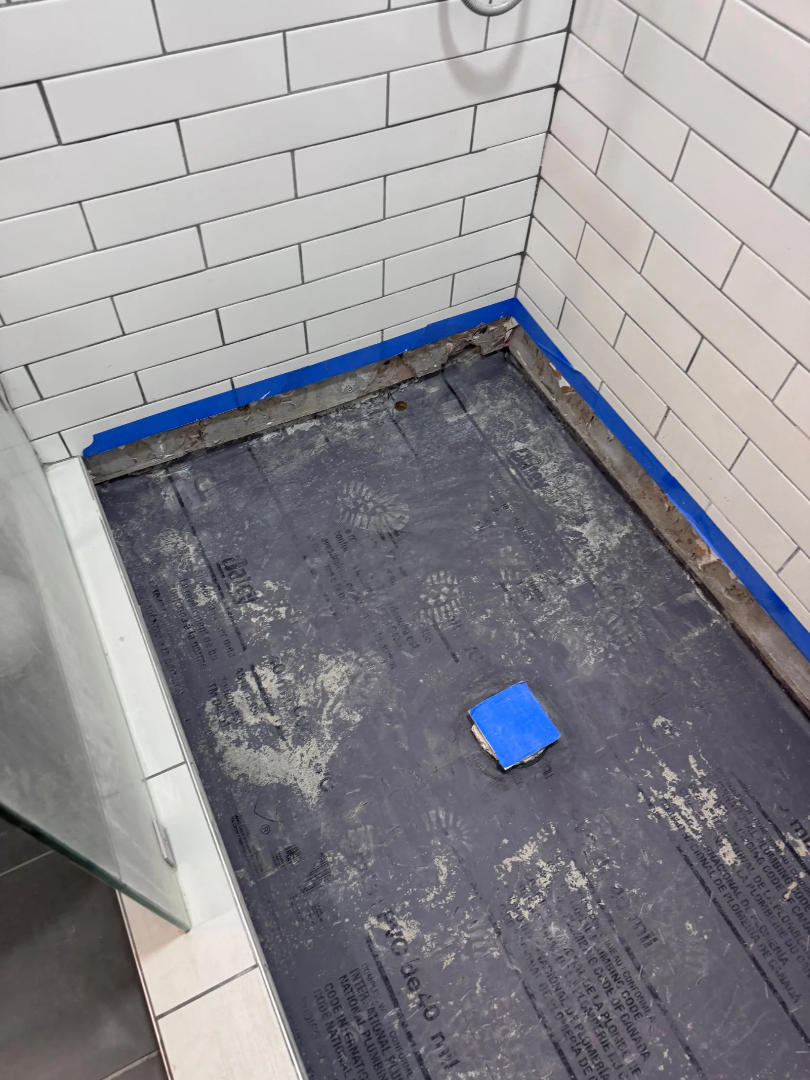 Shower stall under construction with white subway tile walls and blue painter's tape outlining where the shower pan will be i