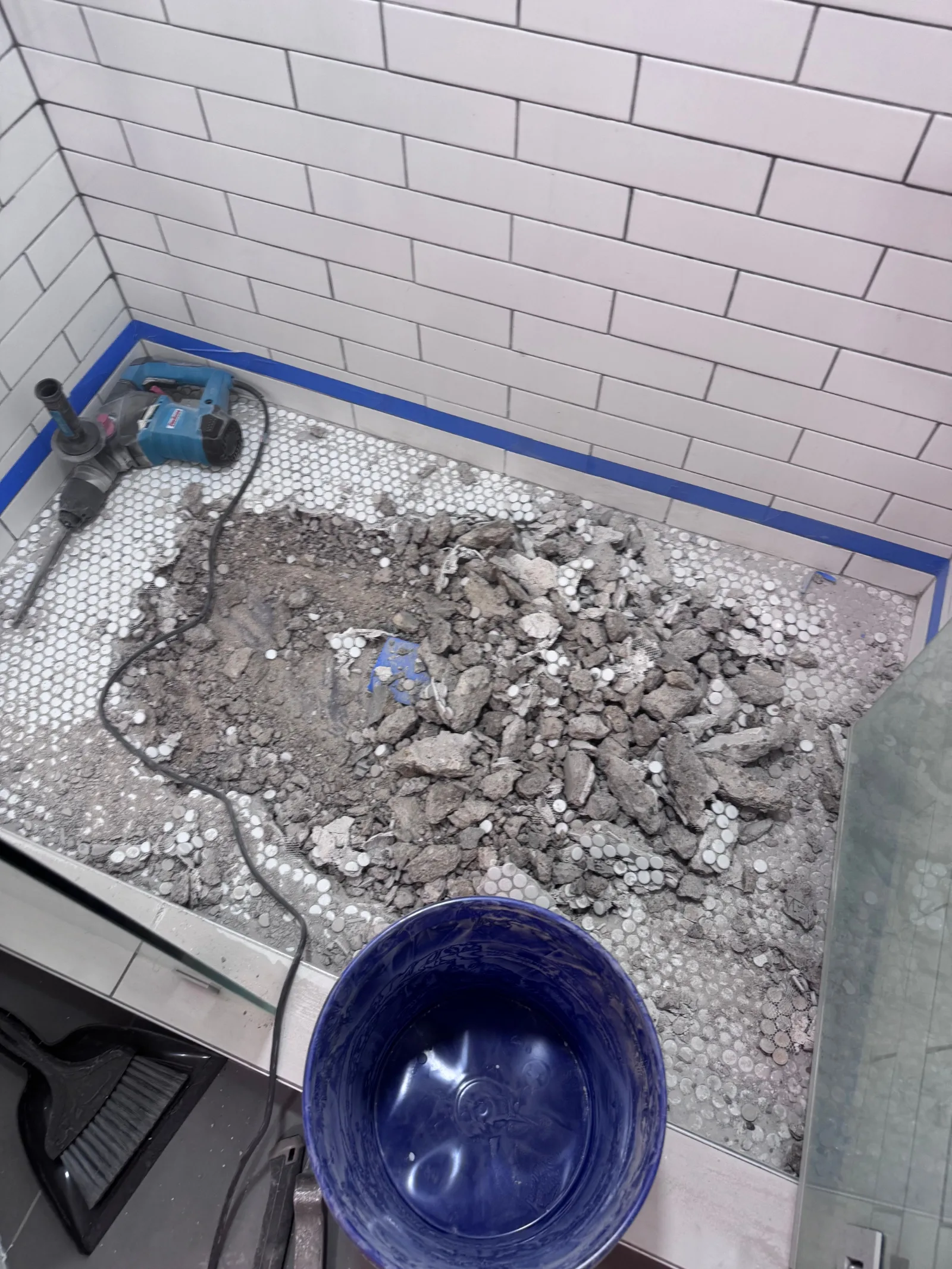 Shower floor with removed tile and concrete debris. White subway tile walls with blue painter's tape along the floor line. A