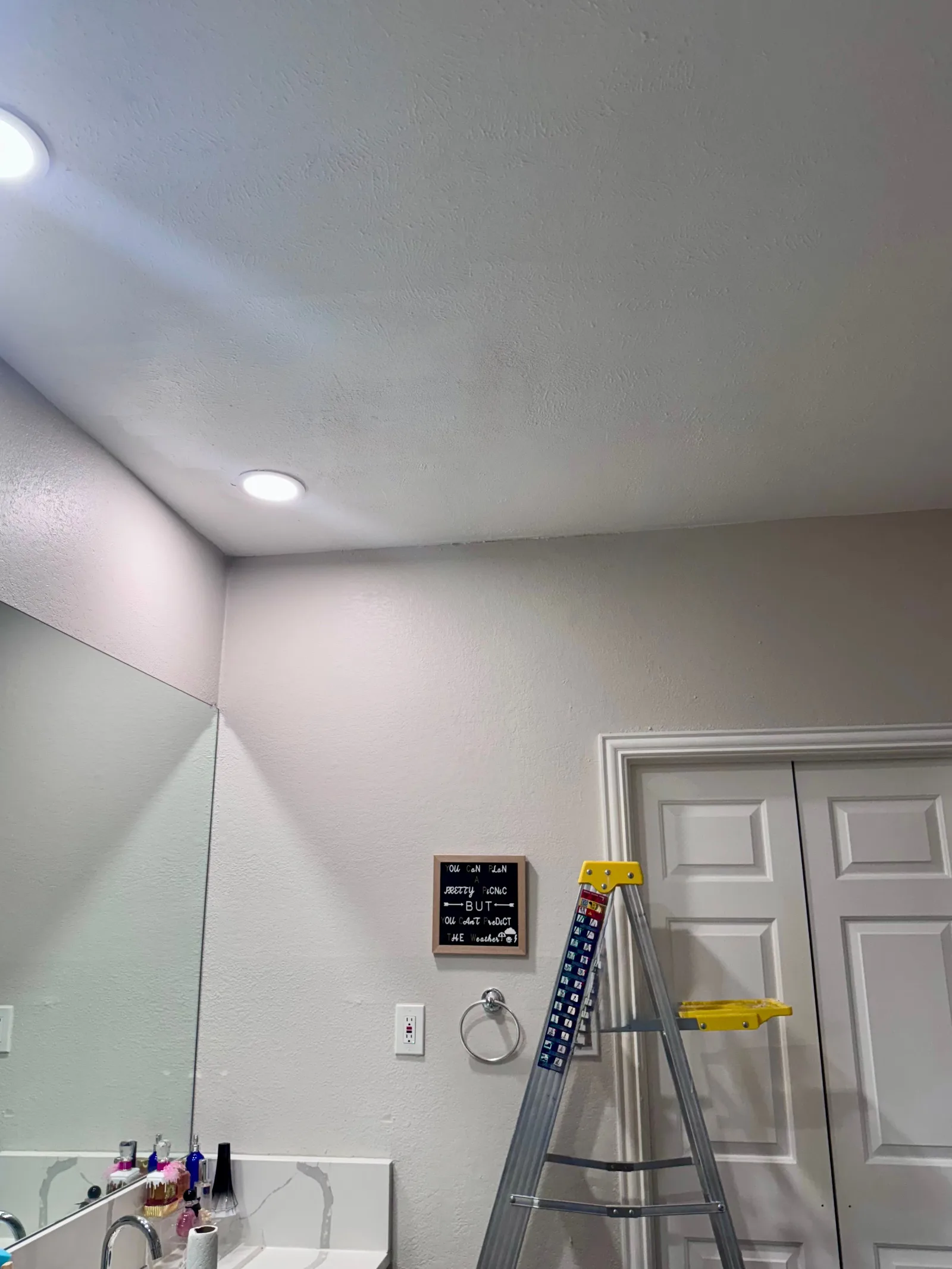 Bathroom with a light beige painted wall and a white textured ceiling with recessed lighting. A mirror, white quartz countert