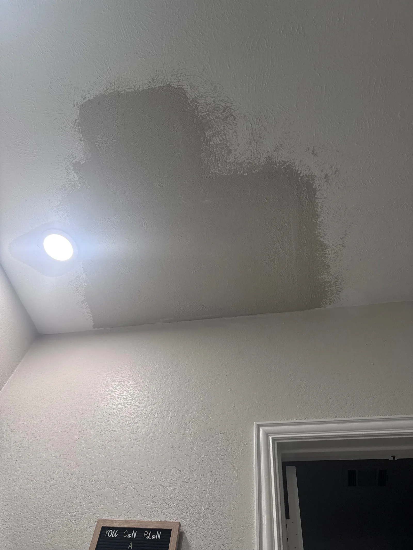 A ceiling with a patchwork of light gray primer over a water-stained area, next to a recessed light fixture.